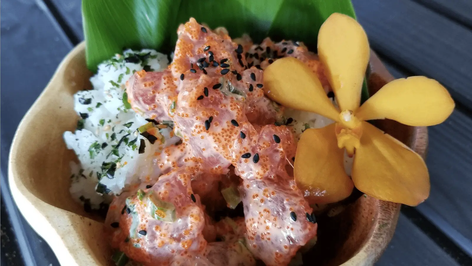 A close-up view of a bowl containing a serving of spicy poke, with pieces of marinated raw fish, rice topped with seaweed and sesame seeds. A yellow flower and a green leaf garnish the dish, adding a touch of color and elegance—just like you'd find in the best places Oahu offers.