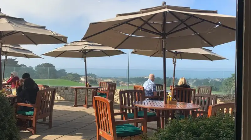 A scenic patio with wooden tables and chairs under large umbrellas, overlooking an expansive view of the ocean and distant hills. Several people are seated at the tables, enjoying the best restaurant view on the Monterey Peninsula under a clear, blue sky.