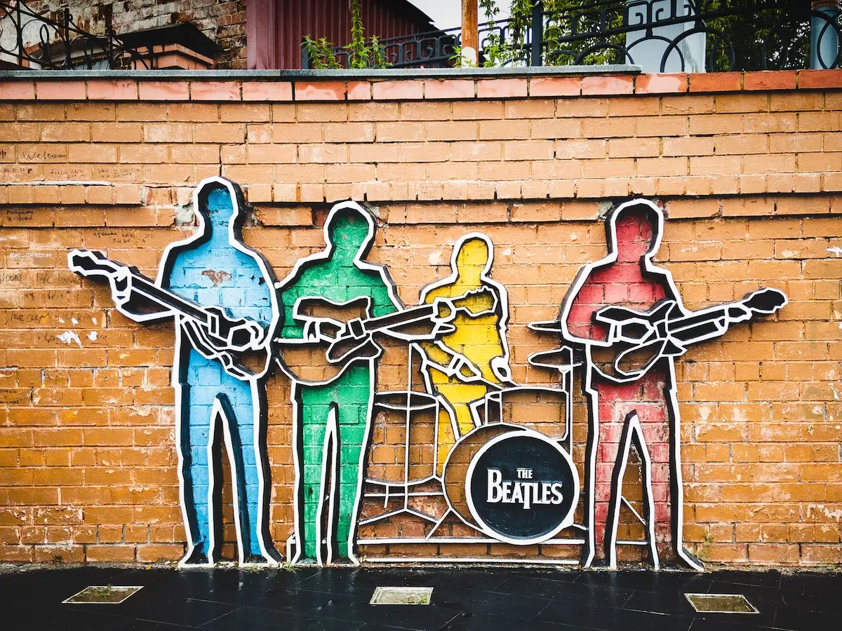 A vibrant mural on a brick wall depicts four colorful, stylized figures of musicians. Each figure holds an instrument, with a drum set displayed in the center. The drum prominently features the name "The Beatles." Experiencing this artwork is one of the best things to do on Monterey Peninsula in November.