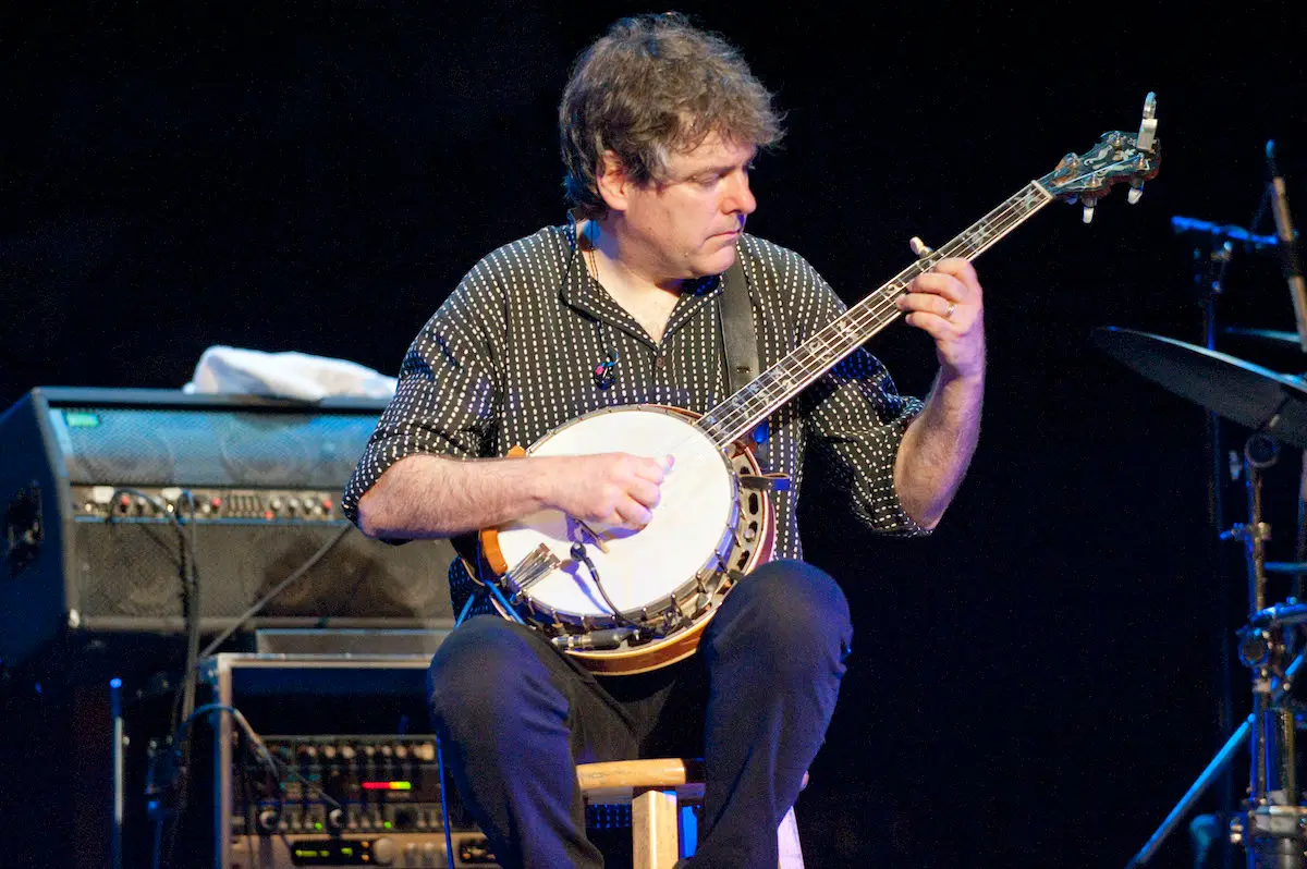 A musician with curly hair sits on a stool on stage, intensely playing a banjo. He wears a black, dotted long-sleeve shirt and black pants. Behind him are various musical equipment and amplifiers. The background is dark, focusing attention on the performer—one of the best things to do Monterey Peninsula November offers.