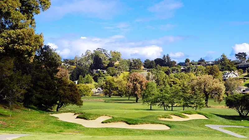 A scenic view of a lush golf course, perfect for golfing, features a well-manicured green surrounded by sand bunkers in the foreground. Mature trees and houses dot the landscape in the background under a blue sky with scattered clouds. It's one of the best golf courses in the greater Bay Area.