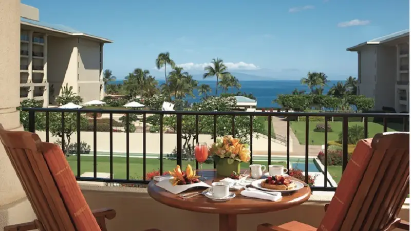 A balcony with a table set for breakfast, including coffee, juice, and pastries, overlooks a resort courtyard with lush greenery and tall palm trees. If you're wondering where to stay in Hawaii, consider this spot—with clear views of the ocean and distant mountain range under a blue sky.