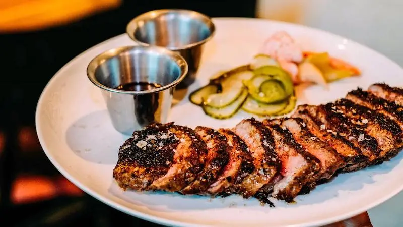 A plate with sliced, seasoned meat garnished with sea salt, accompanied by two small metal cups containing dipping sauces, and a side of pickled cucumber slices. The food is arranged in an appetizing manner on a white plate—perfect for the best happy hour Oahu has to offer.