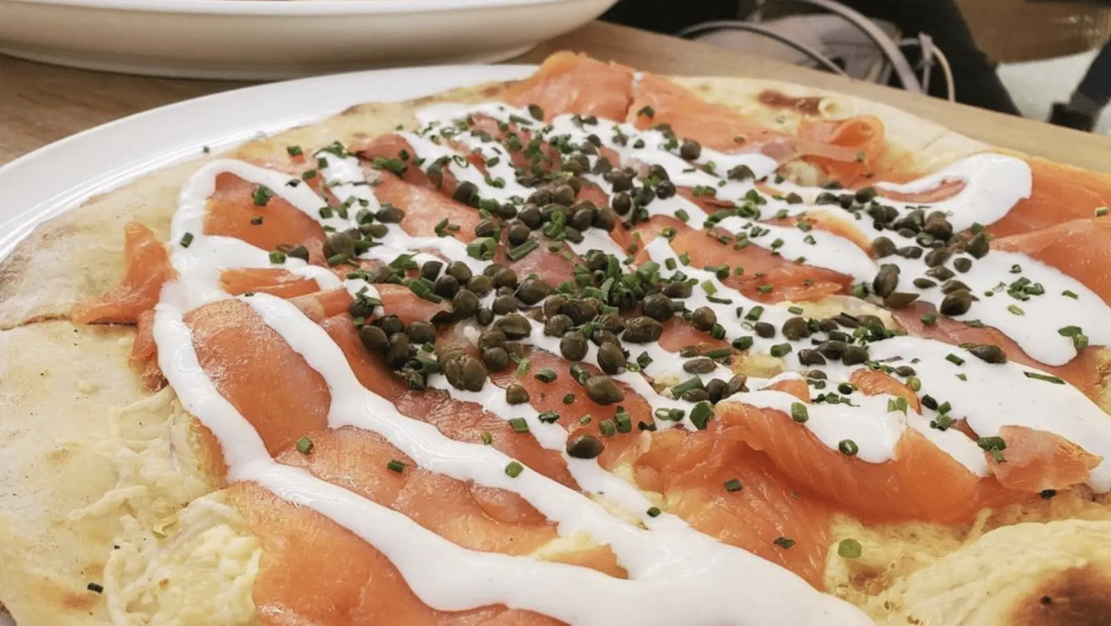 Close-up of a smoked salmon flatbread topped with capers, chopped chives, and drizzled with a creamy white sauce. The flatbread rests on a white plate, offering a glimpse of why this is considered the best breakfast in Napa Valley. Another dish is partially visible in the background.