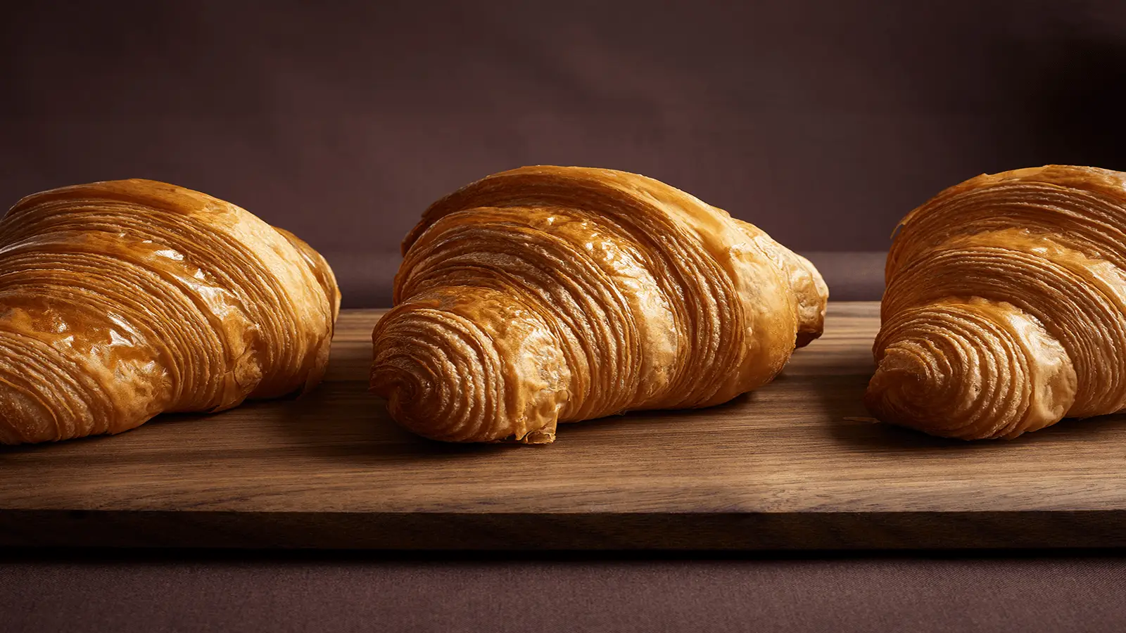 Three golden, flaky croissants are aligned side by side on a wooden board against a dark brown background. Their crisp, layered texture is highlighted by the warm lighting, emphasizing their rich and buttery appearance—truly the best breakfast in Napa Valley.