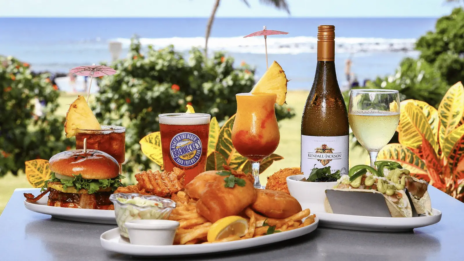 A spread of food and drinks is displayed on a table against a backdrop of the ocean and greenery. The spread includes burgers, fries, fish and chips, and a taco, accompanied by a beer, a tropical cocktail, a bottle of white wine, and a glass of white wine.