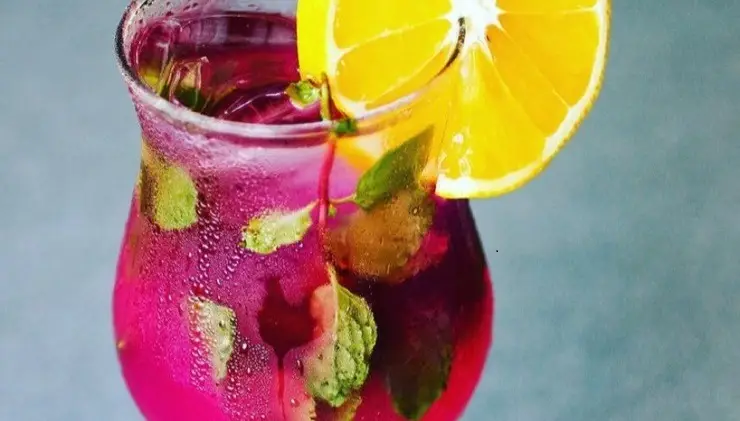 A vibrant pink beverage in a clear glass, adorned with mint leaves and a garnish of a lemon slice on the rim. The drink appears refreshing and is placed on a blue-grey surface, perfect for the best happy hour in Kauai.