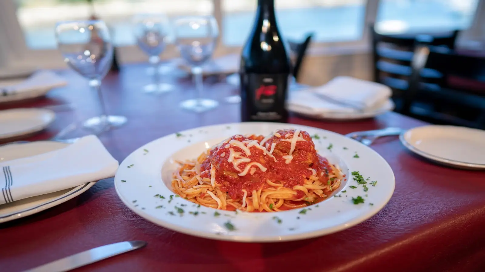 A plate of spaghetti topped with meatballs and marinara sauce garnished with shredded cheese and parsley. The dish is placed on a red tablecloth with folded white napkins, wine glasses, and a bottle of wine in the background, all set against the best restaurant view on the Monterey Peninsula.