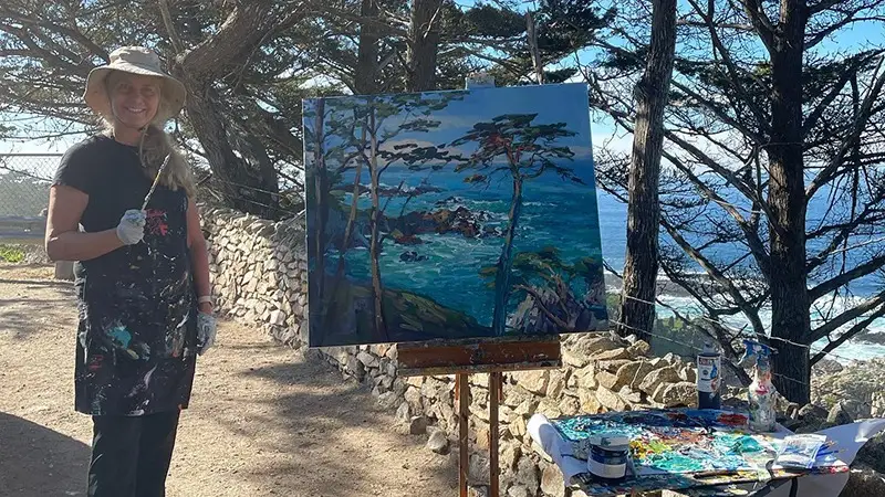 A person, wearing a hat and an apron with paint stains, stands smiling next to an easel holding a painting of a coastal landscape with trees and the ocean. Art supplies are visible on a table beside them, under the serene backdrop of trees and the sea—one of the best things to do on Monterey Peninsula in November.