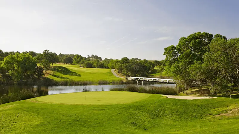 A serene golf course with a well-maintained green in the foreground, bordered by a pond, trees, and a bridge. The lush landscape under a clear blue sky and winding path evoke why this is among the best golf courses Monterey Peninsula offers—one of the must-see sights when exploring Pebble Beach.