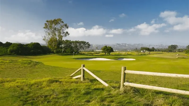 A lush green golf course on a sunny day features a manicured putting green with two sand bunkers. A wooden fence lines the foreground, while trees and mountains are visible in the background under a partly cloudy sky, making it one of the best golf courses on the Monterey Peninsula.