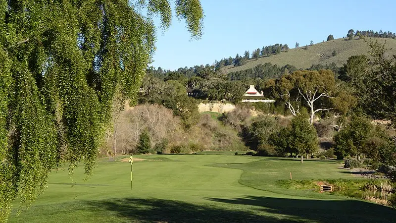 A scenic view of a golf course surrounded by lush greenery and trees, perfect for golfing enthusiasts. The fairway leads to a distant hole marked by a flag. In the background, a hillside is topped with a building, and the sky is clear and blue—one of the best golf courses Monterey Peninsula offers.