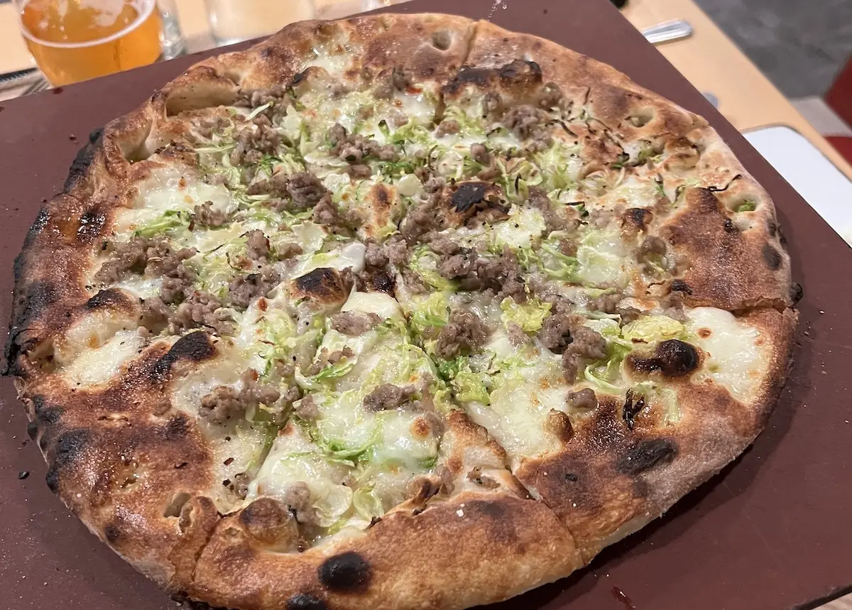 A close-up of a pizza with a thin, crispy crust. The pizza is topped with ground sausage, melted cheese, and shredded green vegetables. The edges of the crust are slightly charred. In the background, there are a few blurred objects, including a glass of beverage at one of the best dinner spots in Sonoma County.