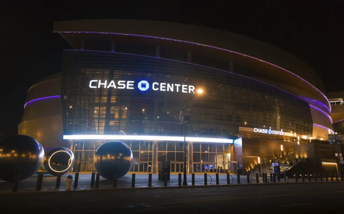 A nighttime view of Chase Center, an arena with illuminated signage and a glass facade. The exterior is lit with purple and white lights. Large decorative spheres are visible in the foreground near the entrance, making it one of the best things to do in San Francisco this May.