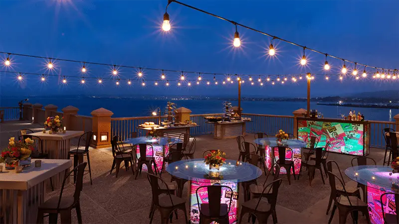 An outdoor restaurant by the sea at dusk, featuring string lights overhead and colorful, illuminated tables. Named the best restaurant view on the Monterey Peninsula, several tables with bouquets of flowers and chairs are set up, with the distant shoreline visible under a clear, deep blue sky.