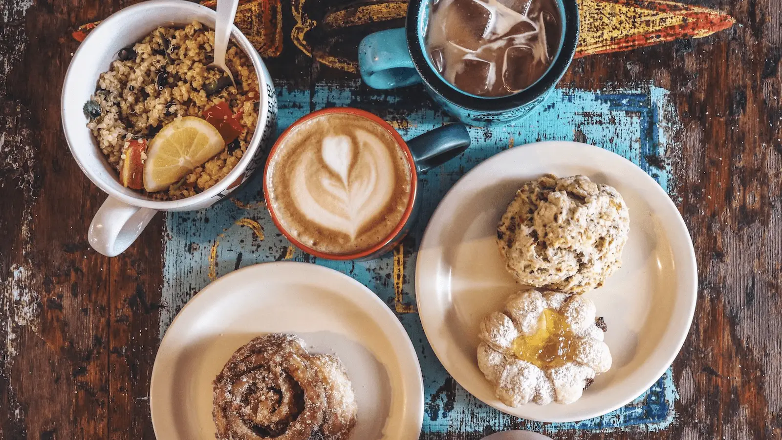 A rustic table features bowls of granola with fresh fruit, a latte with latte art, iced coffee, and plates with a cinnamon roll and two types of scones. The cozy setting on weathered wood and blue painted surfaces makes it the best breakfast spot on the North Shore.