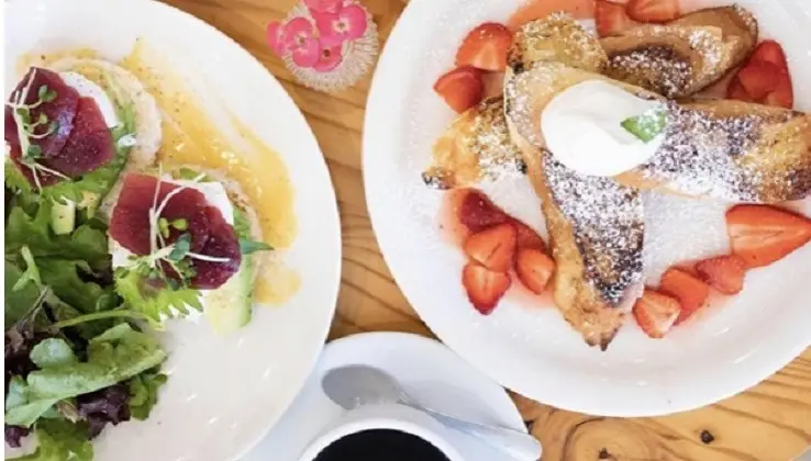 Two plates of delicious brunch food sit on a wooden table at the best breakfast spot in Waikiki. The first plate has poached eggs on greens and toast topped with beets, garnished with microgreens. The second plate features French toast with powdered sugar, fresh strawberries, and a dollop of whipped cream. A cup of coffee is also visible.