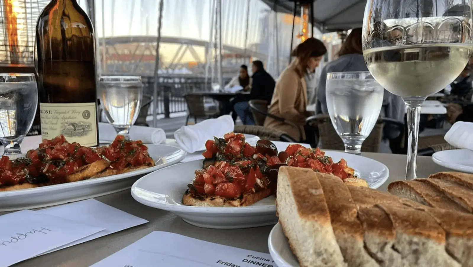 Plates of bruschetta with chopped tomatoes and herbs adorn a table at one of the best dinner spots in Sonoma County. Glasses of water and white wine accompany the dish, with a bottle of wine in the background. Diners enjoy their evening near the railing that overlooks a picturesque bridge and water.