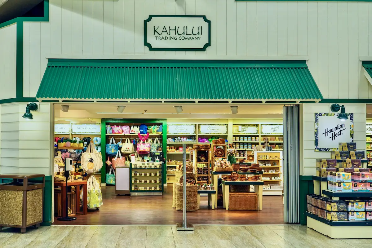A store with a green and white exterior named "Kahului Trading Company." The interior is visible, showing various products like bags, snacks, and souvenirs neatly arranged on shelves and displays. The entrance is open, inviting customers inside to explore while picking up tips on Duty Free Shopping in Maui airport Hawaii.