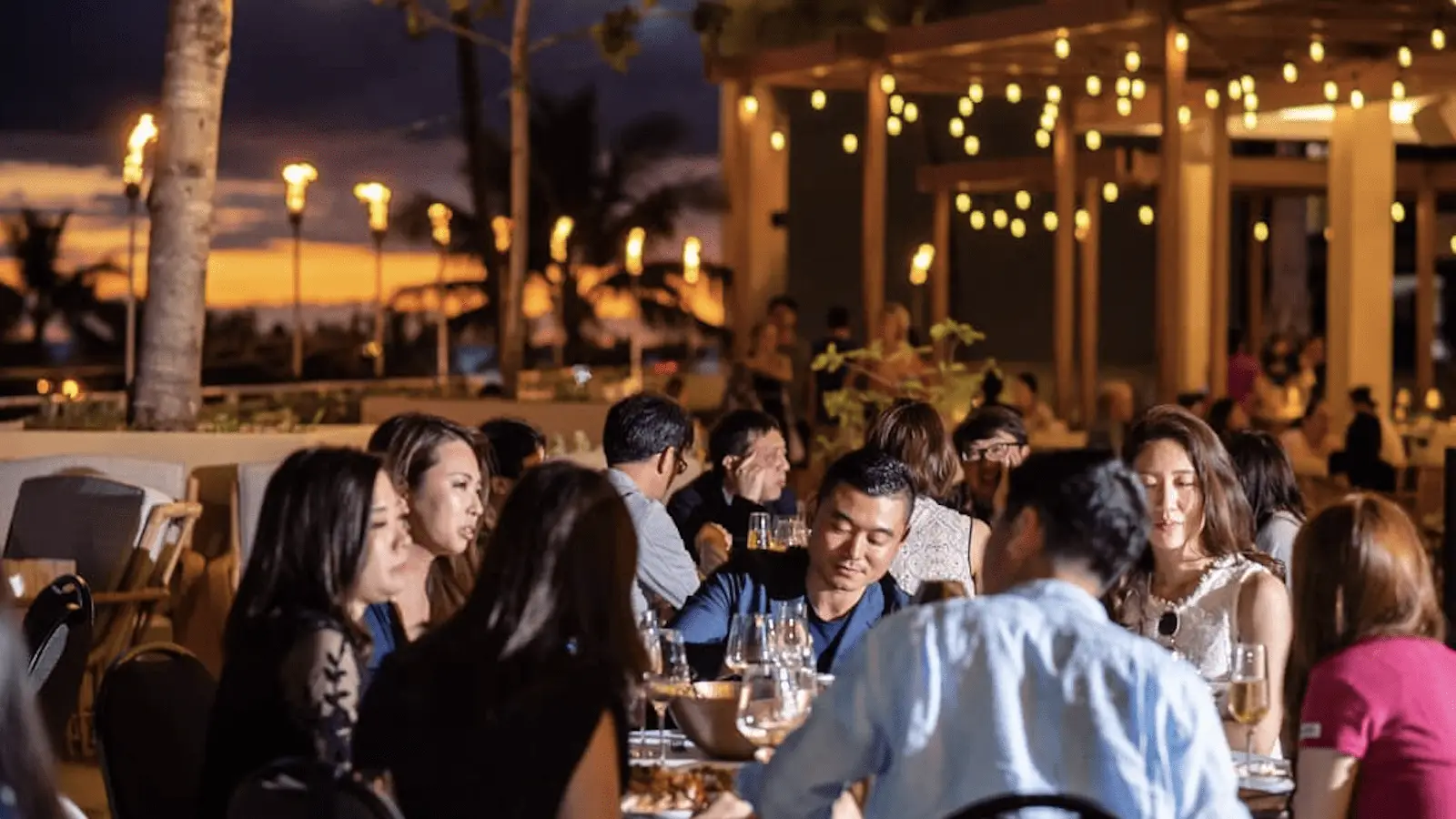 A group of people enjoying a dinner gathering at an outdoor restaurant in the evening, savoring the best sunset dining Oahu has to offer. They are seated around tables under string lights with a beautiful sunset and palm trees visible in the background. The atmosphere is lively and festive.