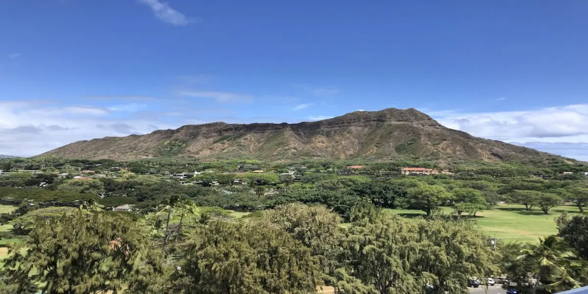 A lush, green landscape stretches out before the rugged volcanic formation of Diamond Head Reservation in Hawaii, under a bright blue sky. Small buildings are scattered among the greenery at the base of the formation, which features a series of ridges and a gently sloping profile.