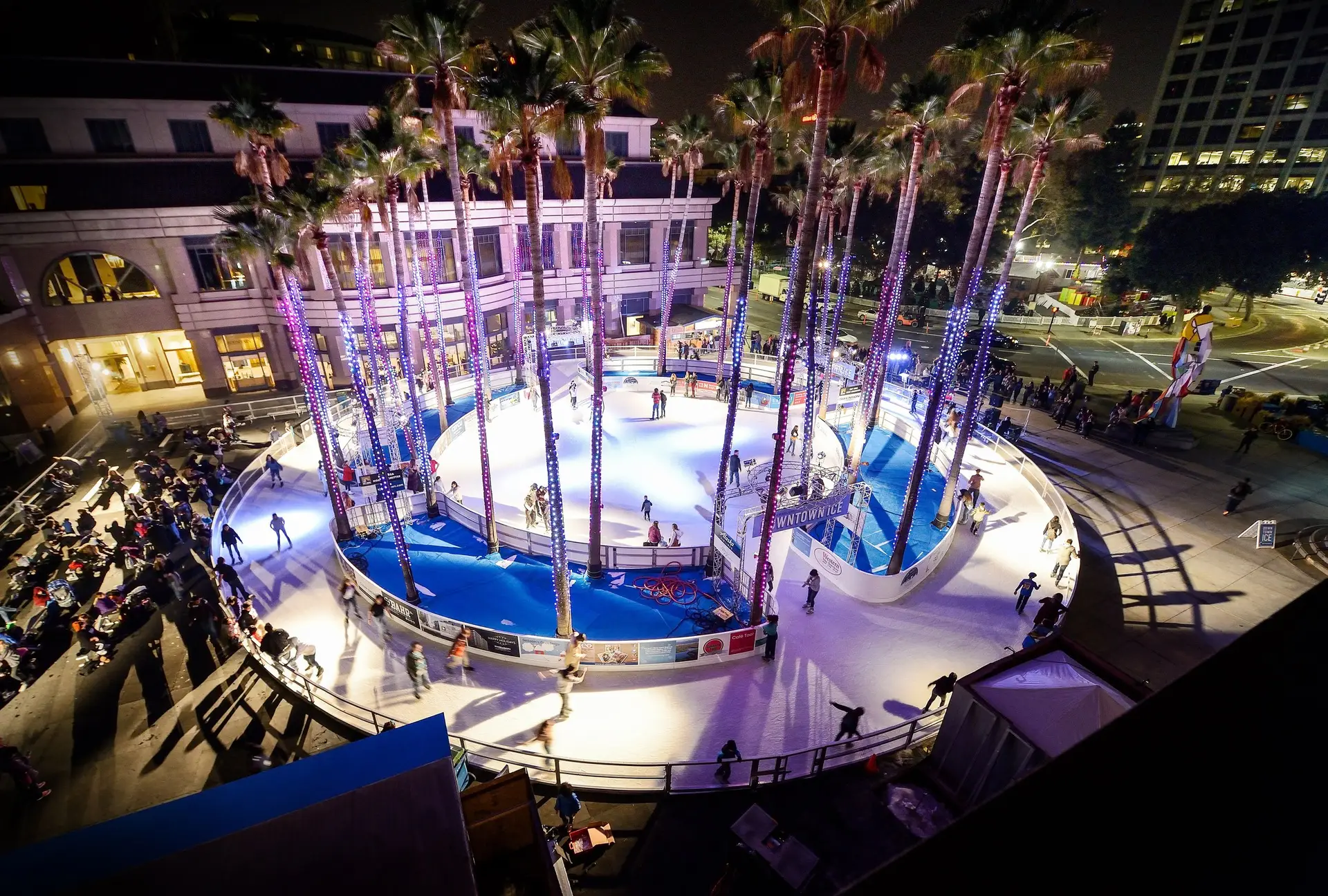 A night view of an outdoor ice skating rink surrounded by tall palm trees wrapped in twinkling lights, skaters glide on the ice while spectators watch. Set in an urban area with buildings and streets illuminated in the background, it’s one of the best things to do South Bay November.