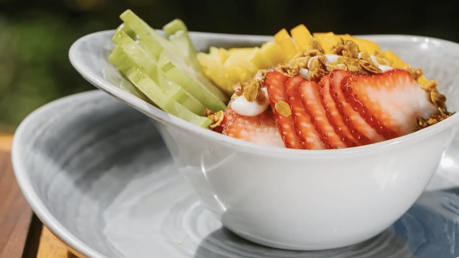A white bowl filled with neatly arranged slices of cucumber, strawberries, and mango, topped with granola and a dollop of yogurt, is placed on a gray plate. The background is blurred with greenery, capturing the essence of the best breakfast Kaanapali has to offer.
