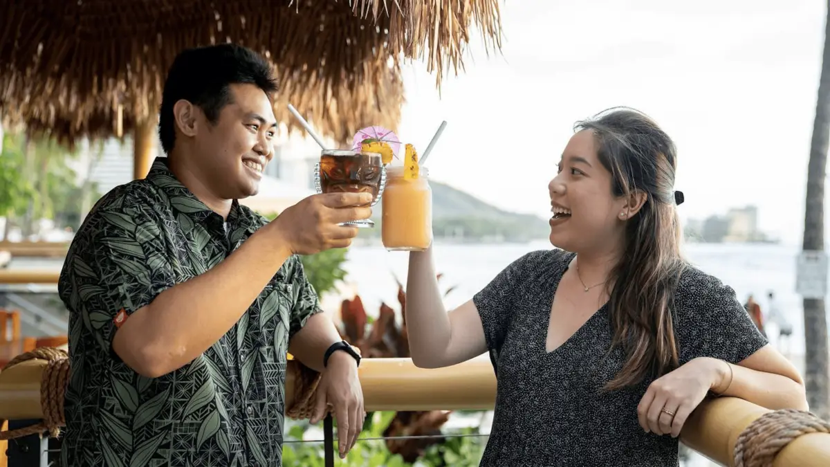 A man and a woman smiling and toasting with drinks under a thatched roof, enjoying the best sunset dining Oahu offers. The man holds a dark beverage with a lemon garnish, while the woman sips on a yellow, blended drink with a straw and fruit garnish. Lush greenery and a body of water serve as their serene backdrop.