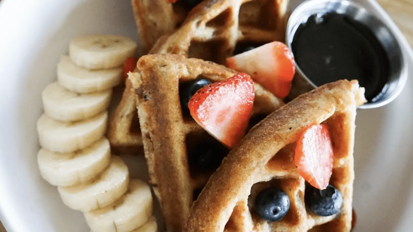 A plate of waffles topped with sliced strawberries and blueberries, considered the best breakfast on the North Shore. To the side, there are several slices of banana lined up neatly. A small metal container of syrup is placed in the upper right corner of the plate.
