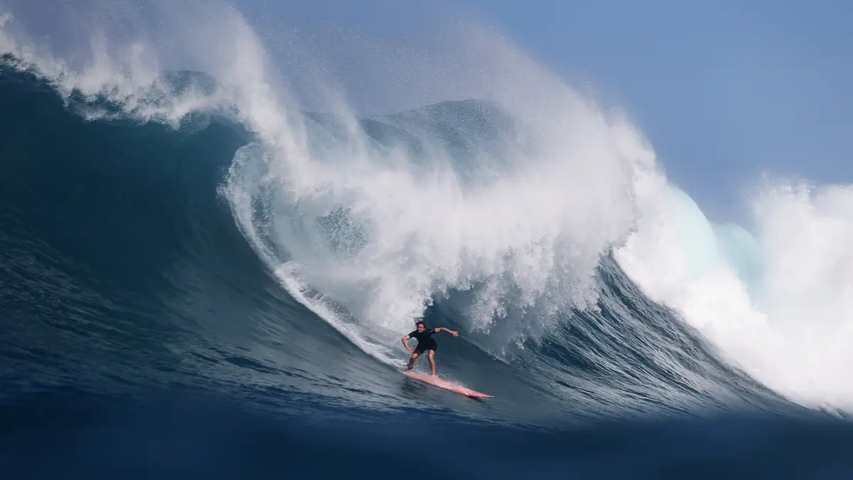 A surfer skillfully rides a large, powerful wave with a dramatic curl at the Eddie Aikau Invitational. The ocean spray and blue sky add to the dynamic intensity of the scene, showcasing the thrill and skill involved in surfing such challenging waves.