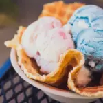 A delicious serving of ice cream in a waffle bowl, hailed as one of the best desserts North Shore. The bowl contains three scoops of ice cream in pastel colors—blue, pink, and white. The crispy waffle contrasts with the creamy texture of the ice cream, set on a metal patio table with a blurred background.