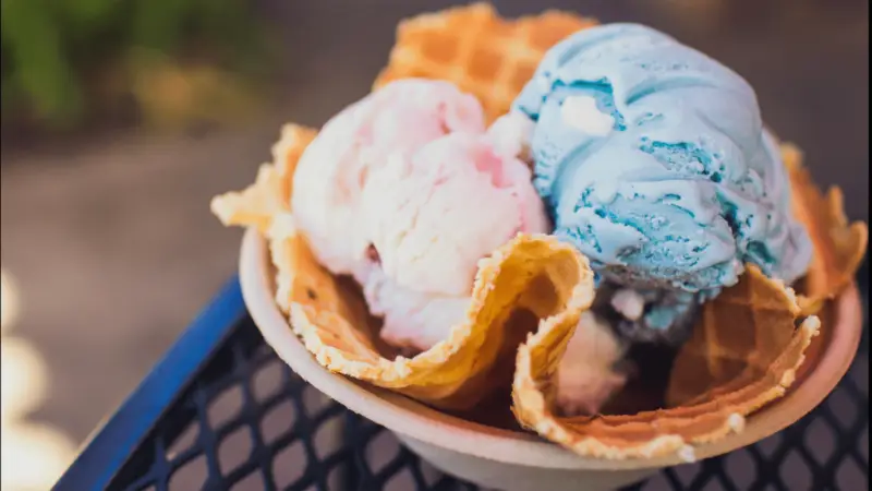 A delicious serving of ice cream in a waffle bowl, hailed as one of the best desserts North Shore. The bowl contains three scoops of ice cream in pastel colors—blue, pink, and white. The crispy waffle contrasts with the creamy texture of the ice cream, set on a metal patio table with a blurred background.