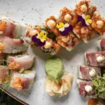 A plate of assorted sushi rolls, perfect for the best dinner in Waikiki, presented in a visually appealing arrangement. Three varieties are garnished with sauces, microgreens, and edible flowers. A serving of pickled ginger and a wasabi dollop are placed in the center. The white plate is on a dark surface.