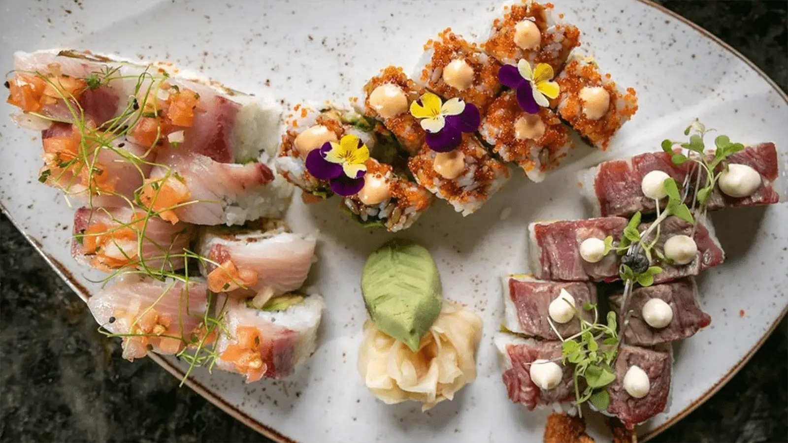 A plate of assorted sushi rolls, perfect for the best dinner in Waikiki, presented in a visually appealing arrangement. Three varieties are garnished with sauces, microgreens, and edible flowers. A serving of pickled ginger and a wasabi dollop are placed in the center. The white plate is on a dark surface.