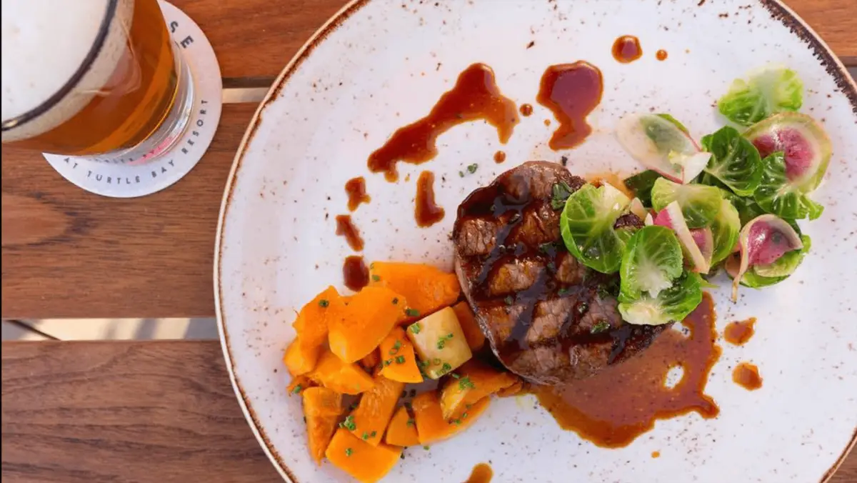 A plate of steak adorned with a rich sauce, accompanied by cubes of butternut squash garnished with chives, and a side of brussels sprout leaves and thinly sliced radishes. A pint of beer rests on a coaster to the left, all set on a wooden table—truly the best dinner on the North Shore.