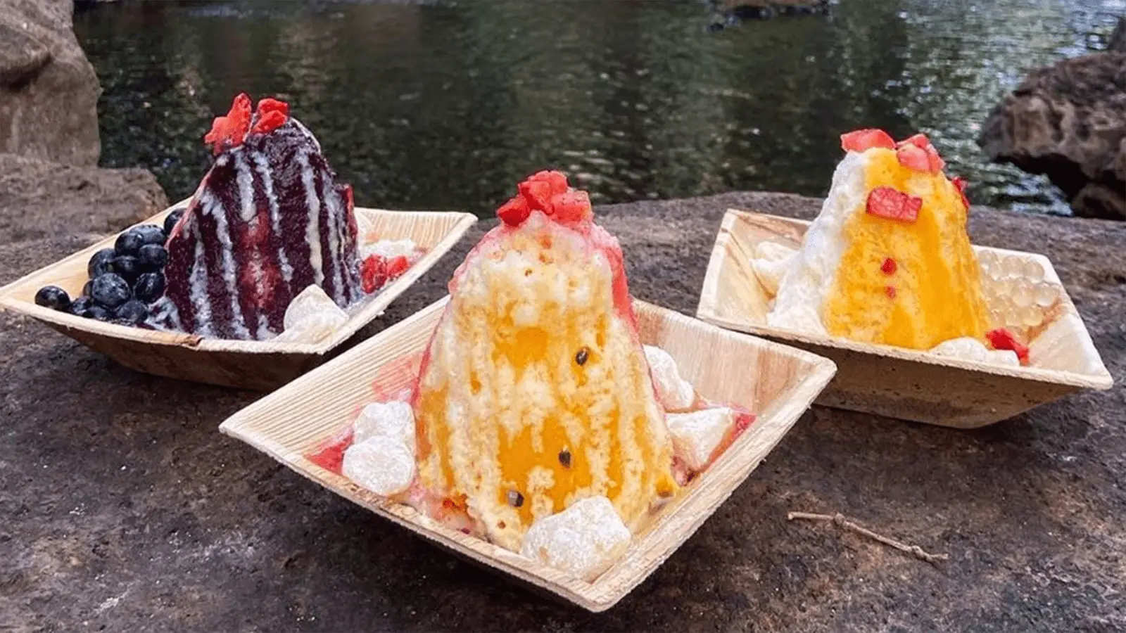 Three plates of the best shave ice in Waikiki topped with different syrups and fruit are set on a stone surface by a lake. The left plate has a dark purple syrup with blueberries, the middle one has yellow syrup with diced red fruit, and the right one has an orange syrup with diced fruit.