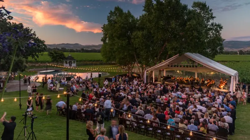 A large outdoor event with a crowd seated under a clear tent canopy. String lights are strung overhead, and a symphony orchestra is performing on stage. The surrounding landscape includes a vineyard, trees, and a beautiful sunset sky in the background—a perfect scene for local getaways.