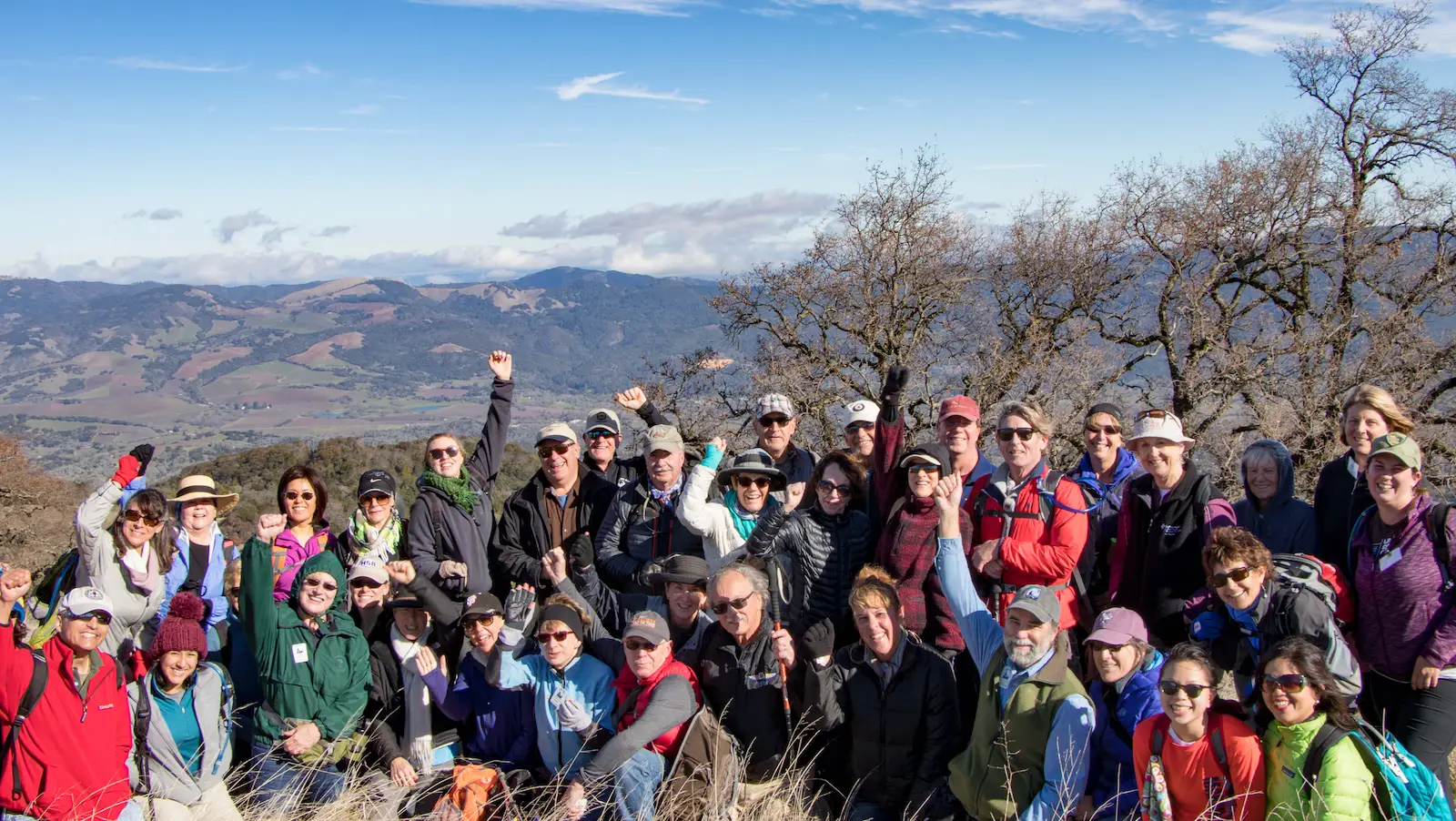A group of approximately thirty people is gathered outdoors on a grassy hilltop in Sonoma, posing for a photo. Dressed in colorful outdoor attire, they smile and cheer with arms raised. The background features a scenic view of rolling hills, valleys, and a partly cloudy sky—one of the best things to do in January.