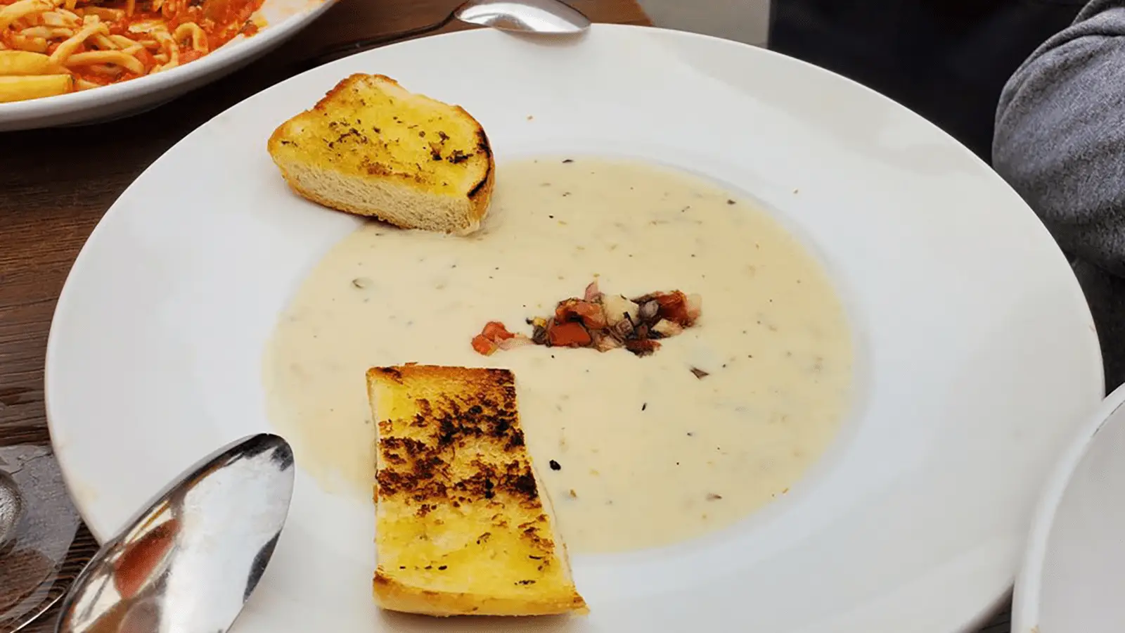 A white plate with creamy clam chowder, hailed as the best on the Monterey Peninsula, garnished with diced vegetables at the center. The chowder is accompanied by two slices of toasted garlic bread on the rim of the plate. A spoon rests on the edge, and pasta is partially visible in the background.