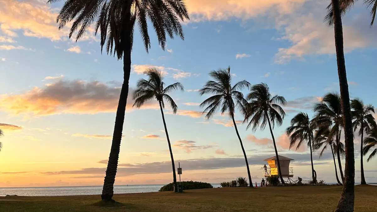 A scenic beach at sunset, with tall palm trees silhouetted against a colorful sky of pink, orange, and blue hues. A lifeguard tower stands near the beach with a few people around it. The tranquil ocean in the background makes this one of the best places to visit on Oahu.