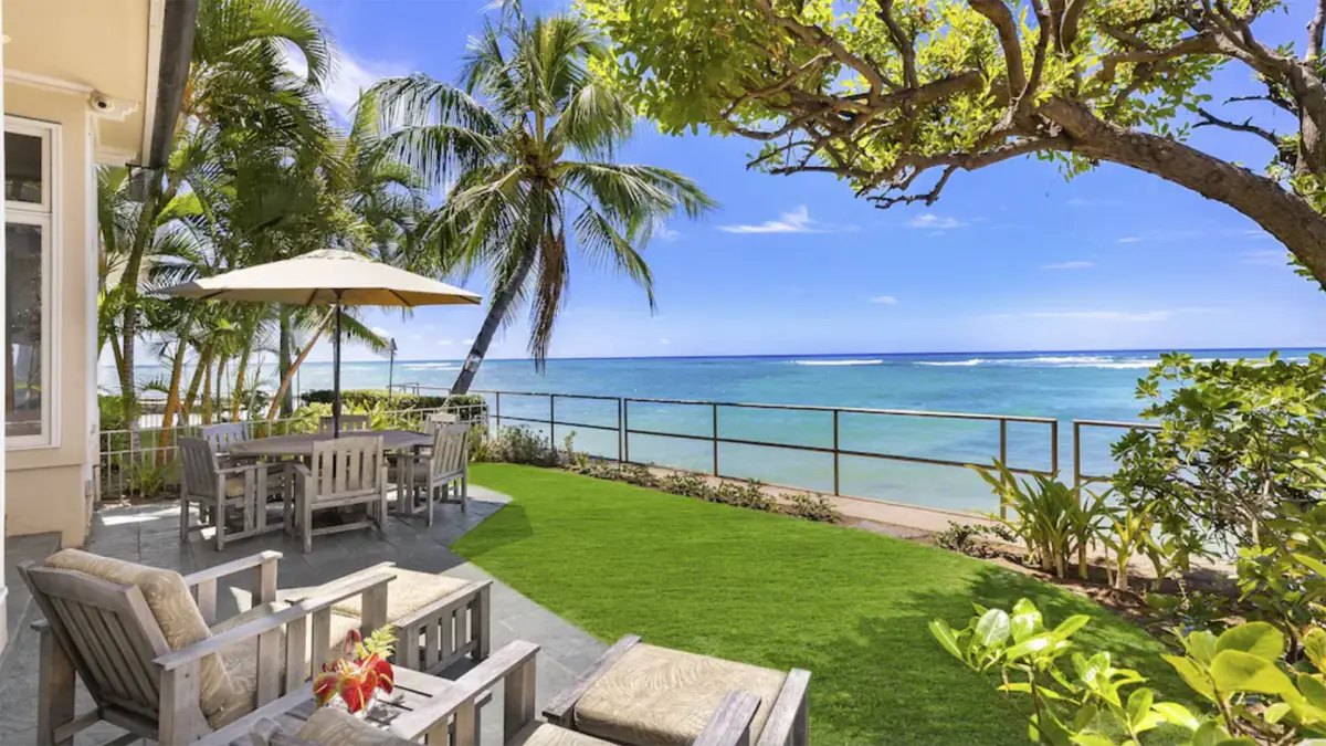 A serene oceanfront patio, part of our exclusive vacation rentals in Hawaii, features wooden outdoor furniture including tables and chairs arranged under a large sun umbrella on a neatly manicured lawn. Palm trees and lush greenery frame the scene, offering a view of the turquoise sea and clear blue sky.