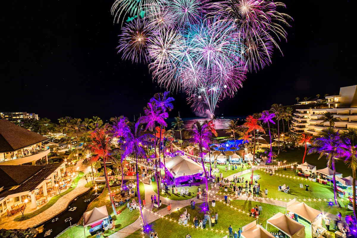 A vibrant night scene with multicolored fireworks exploding in the dark sky above a lively outdoor event. Brightly lit tents, palm trees, and numerous people are gathered on a spacious lawn near the waterfront at the Hawaii Food and Wine Festival, creating a festive atmosphere.