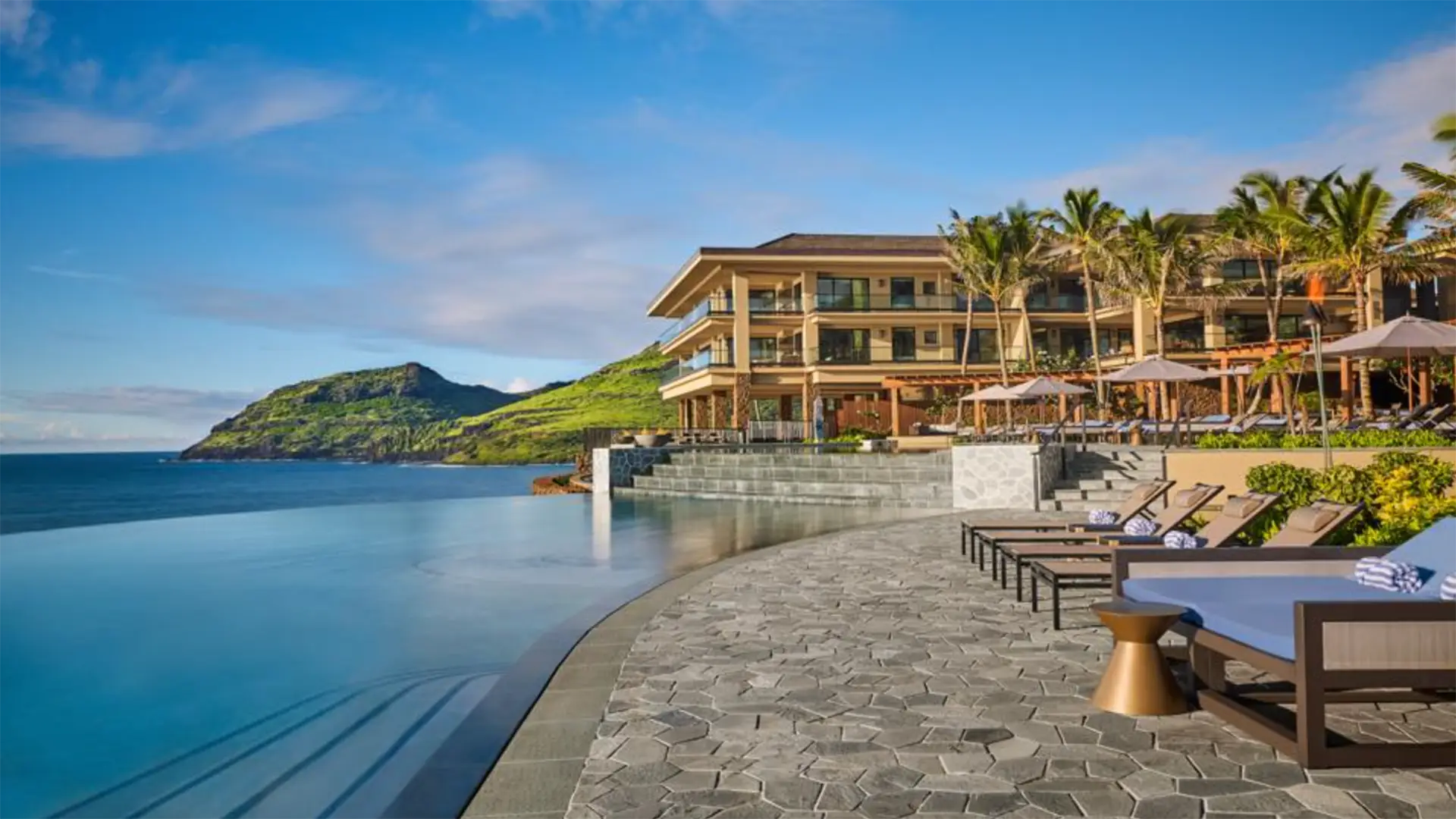 A coastal luxury resort, Timbers Resort Hokuala, features a curved stone pathway leading to an infinity pool overlooking the ocean. Lounge chairs and umbrellas are arranged along the poolside, while a multi-story building stands in the background with palm trees and green hills nearby.