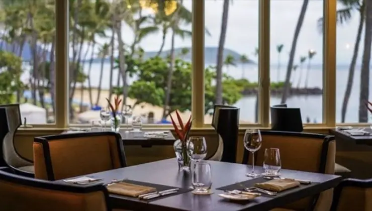 A calming, tropical-themed restaurant scene with neatly set tables and chairs. Large windows offer a scenic view of palm trees, a sandy beach, and the ocean in the background. Experience the best sunset dining in Oahu with folded napkins, glassware, and a small floral centerpiece gracing each table.