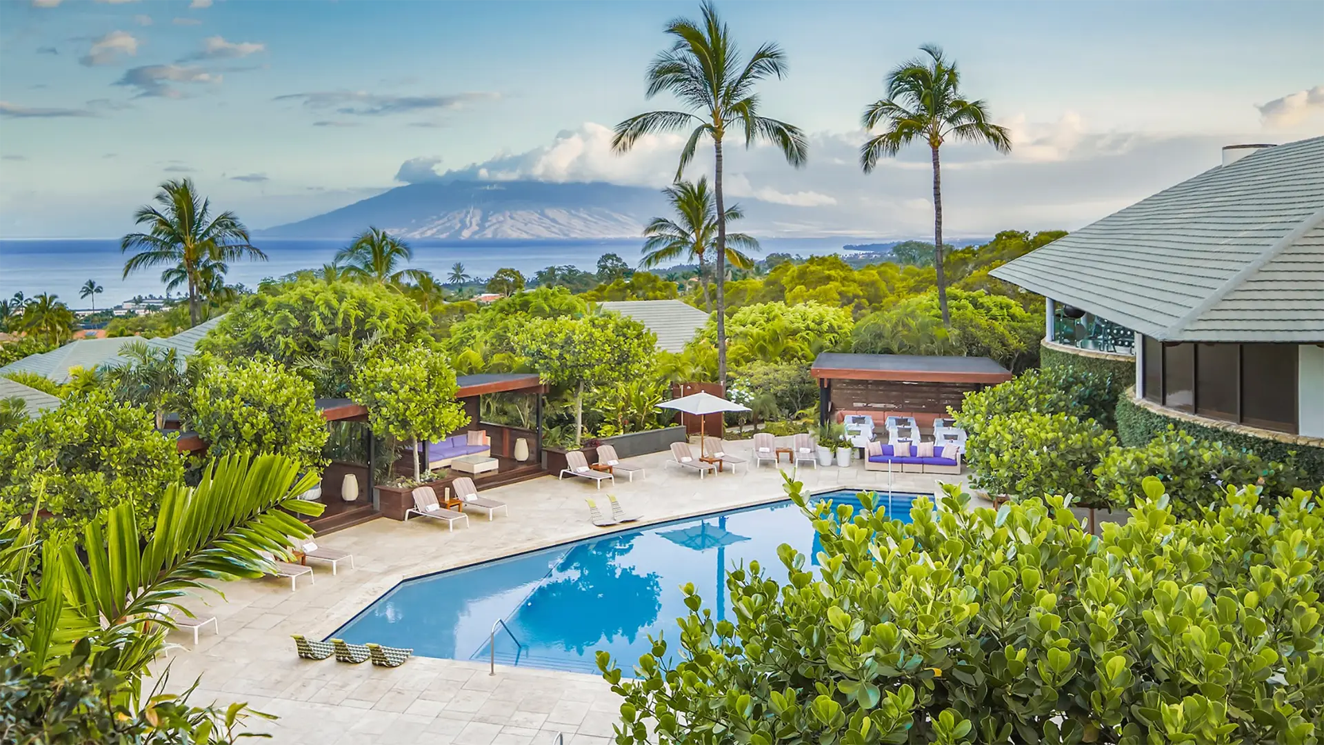 A luxurious outdoor pool area surrounded by lush greenery and palm trees, with lounge chairs and cabanas. In the distance, the ocean water stretches to meet a mountainous island under a partly cloudy sky, creating a serene and tropical setting at one of the best romantic hotels South Maui has to offer.