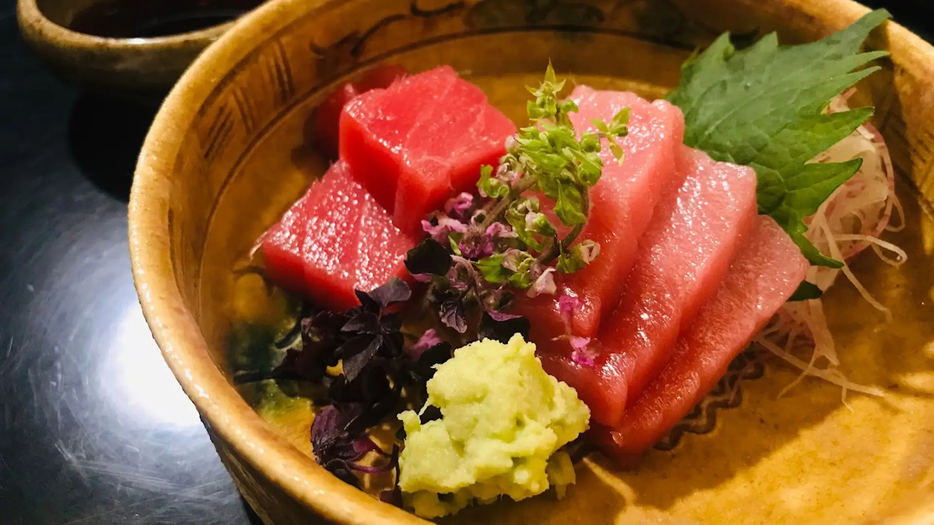 A close-up of a wooden bowl containing sushi-grade slices of raw tuna, garnished with a small mound of wasabi and microgreens, and accompanied by a dark green leaf and shredded white radish. The bright red tuna is neatly presented. Experience the best sushi Kauai offers in every bite.