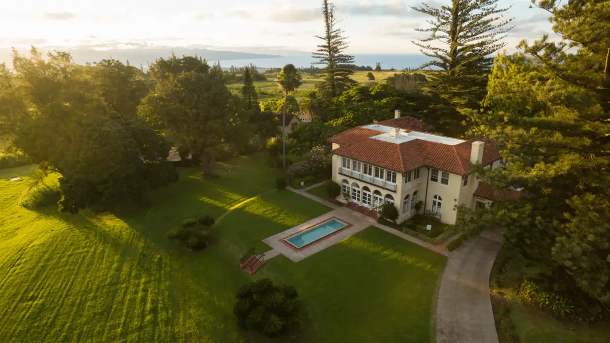 Aerial view of a large, two-story house with a red-tiled roof, surrounded by lush greenery and tall trees. The house features numerous windows and an outdoor rectangular pool. A paved driveway leads to the entrance, reminiscent of the scenic landscapes you find in popular places to visit on Maui.