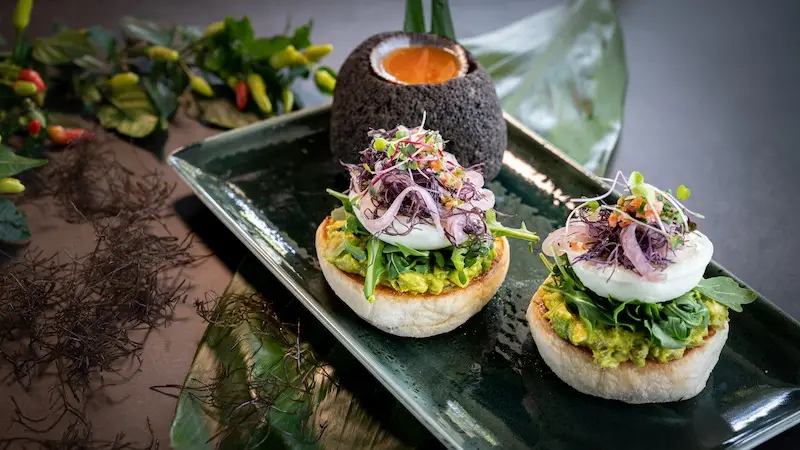 A rectangular plate holds two gourmet avocado toast portions topped with poached eggs, microgreens, and purple sprouts. Behind them is a round stone bowl filled with an orange dip or sauce, reminiscent of the vibrant flavors found in Maui. Arranged greenery adds to the presentation.