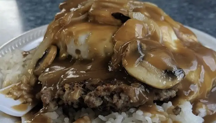 A plate of Loco Moco shows a hamburger patty topped with a fried egg, brown gravy, and sliced mushrooms, all served over a bed of white rice. The background features a blueish surface, suggesting an indoor dining setting. This dish could easily be the best breakfast on the North Shore.
