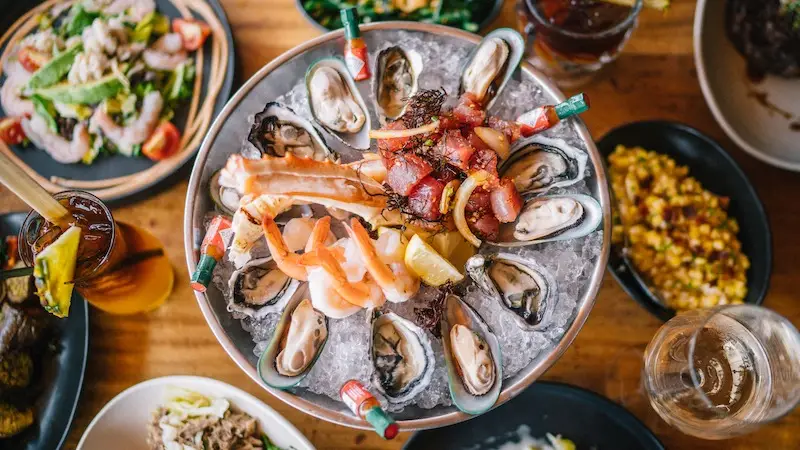 A top-down view of a seafood platter consisting of oysters on ice, garnished with lemon wedges, and accompanied by various appetizers and sides including salads and mixed vegetables. A glass of white wine and other drinks can be seen on the wooden table, perfect for the best Sunset Dining in Oahu.